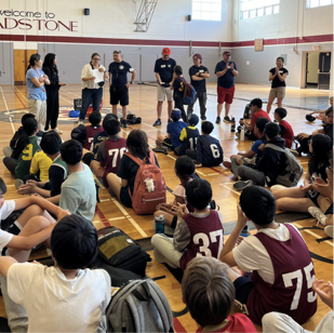 Group of youth seated on gym floor listening to adults standing and speaking in front of them in a basketball court.