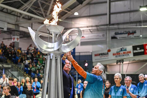Two people light a large torch inside a sports arena filled with seated spectators.