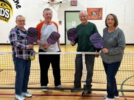 Four adults holding pickleball paddles standing behind a net in an indoor gymnasium.