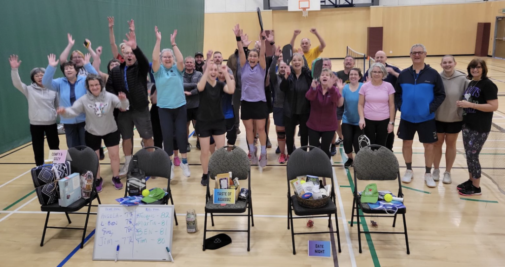 Group of people in a gymnasium cheering behind chairs with gift baskets and whiteboards showing pickleball scores.