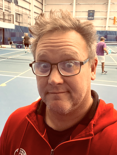 Man with glasses and messy hair wearing a red jacket standing inside a gymnasium with pickleball courts in the background