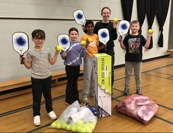 Five children and one adult standing in a gym holding pickleball paddles and balls, with bags of equipment on the floor.