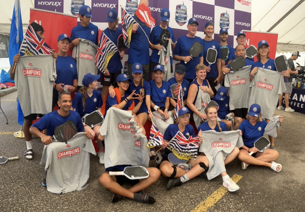 Group of young athletes in blue uniforms holding championship shirts and trophies under a tent at a sports event