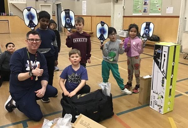 Group of children and an adult in a gym holding pickleball paddles and standing near equipment boxes and bags