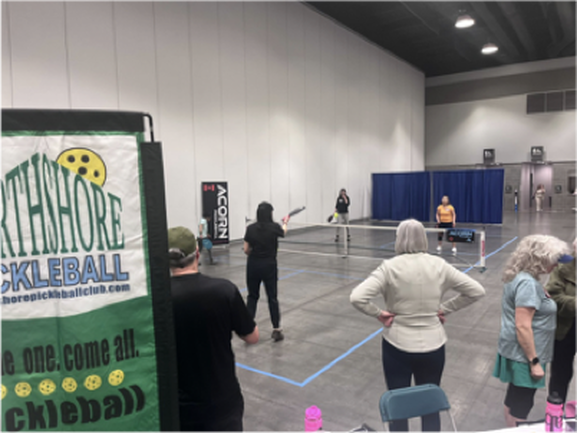 Indoor pickleball game with players and spectators in a large convention hall with blue curtains and gray walls.