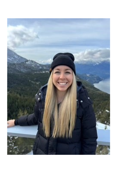 Smiling woman with long blonde hair wearing a black jacket and beanie standing outdoors with mountains and trees in the
