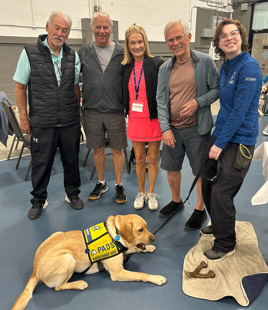 Five people standing indoors on a gym floor with a yellow service dog lying down wearing a PADS vest.