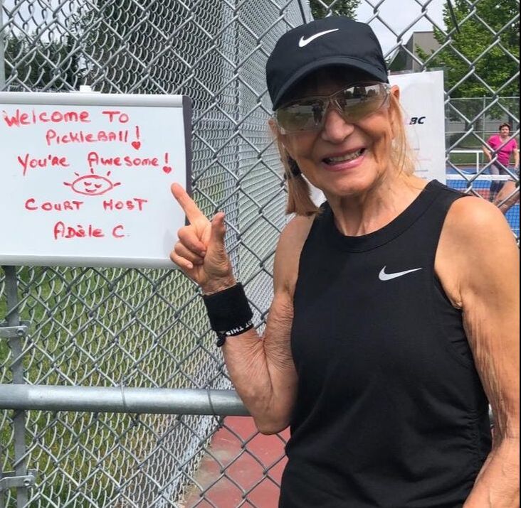 Smiling woman in black Nike cap and shirt points to a handwritten sign welcoming people to pickleball.