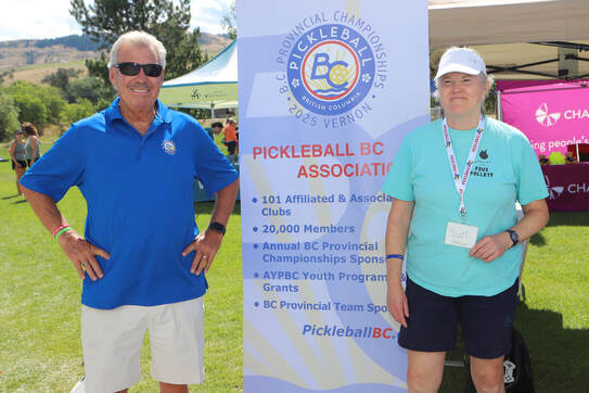 Two men standing outdoors beside a Pickleball BC Association banner at a sunny event with tents and hills in the background.