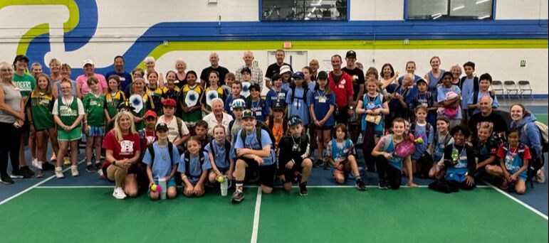 Large group of children and adults posing on an indoor pickleball court with colorful wall mural behind them