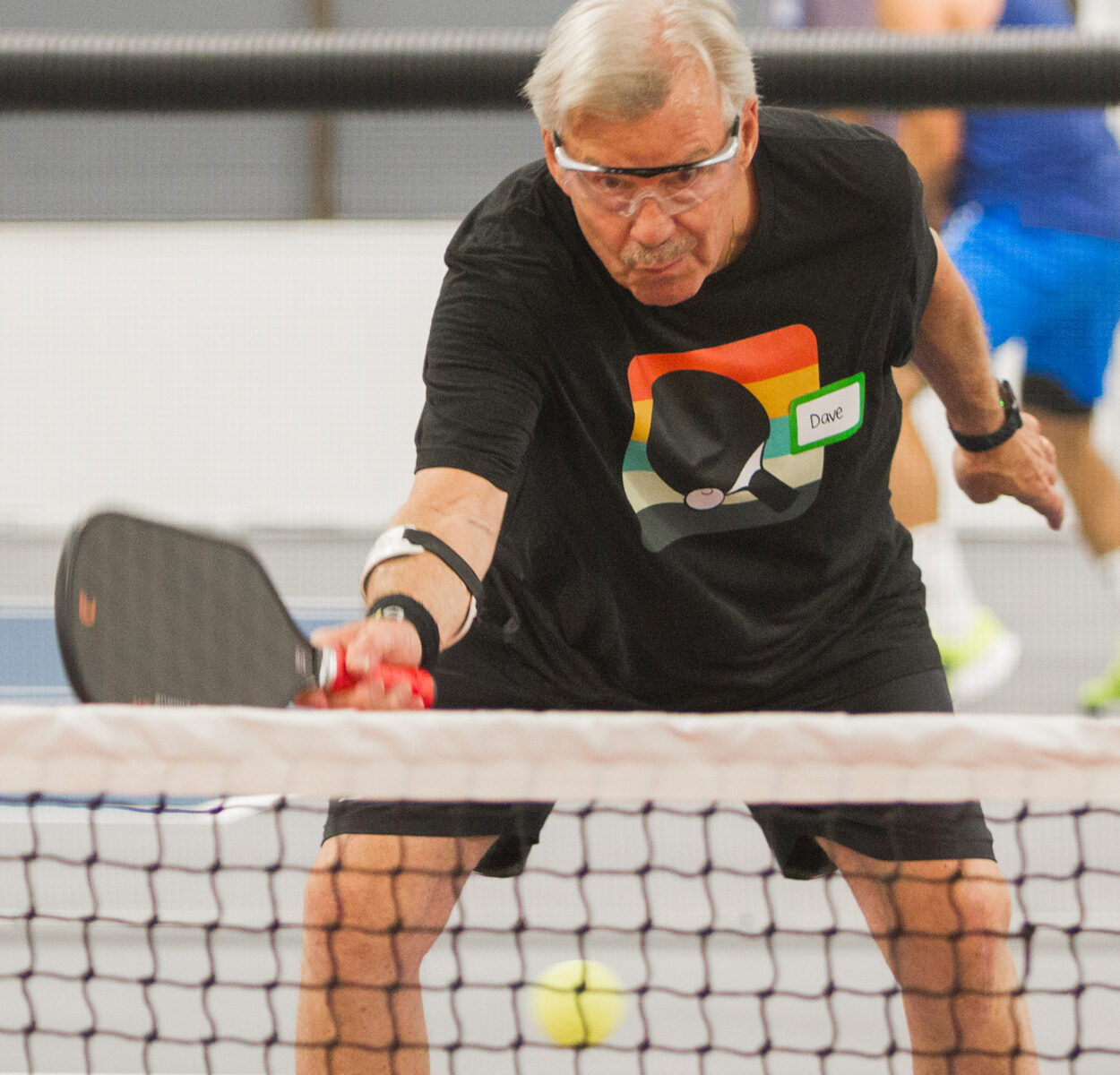 Older man wearing glasses and a black shirt with a paddle graphic, reaching to hit a pickleball near the net