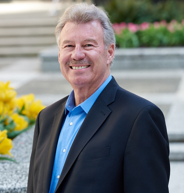 Middle-aged man with gray hair wearing a blue shirt and dark blazer, smiling outdoors near yellow flowers and steps
