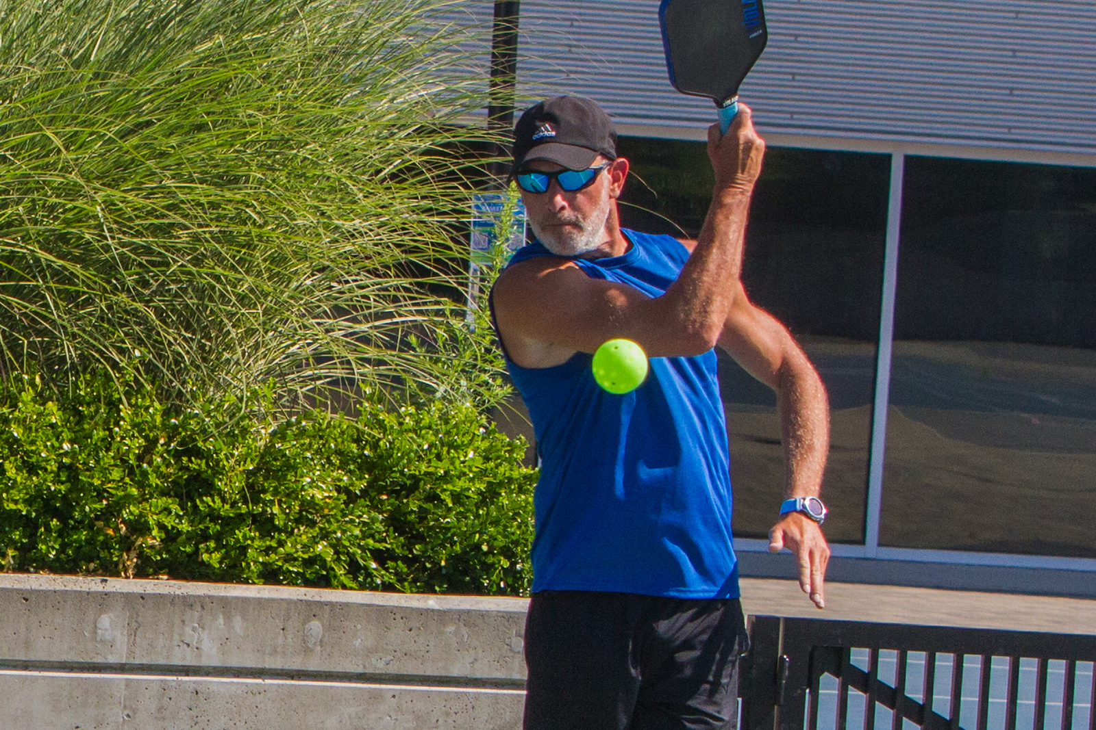 Man in blue sleeveless shirt and black cap hitting a yellow pickleball with a paddle outdoors near bushes and a building