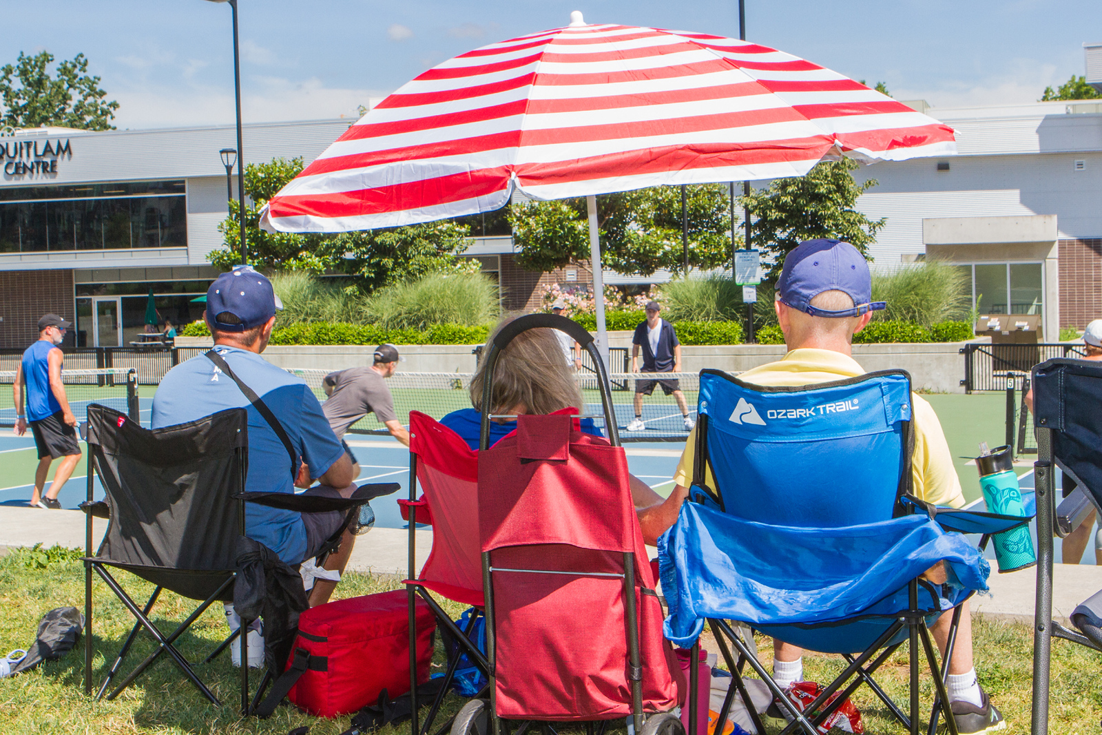 Three people seated in lawn chairs under a red and white striped umbrella watching a pickleball game.