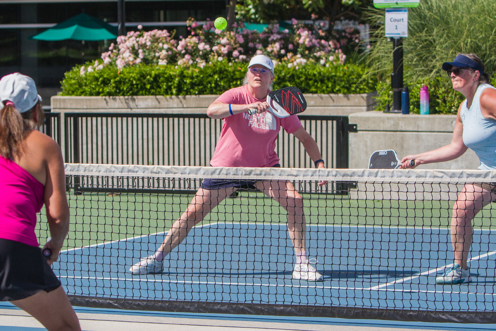 Four women playing pickleball on an outdoor court, one in pink shirt hitting a ball over the net.