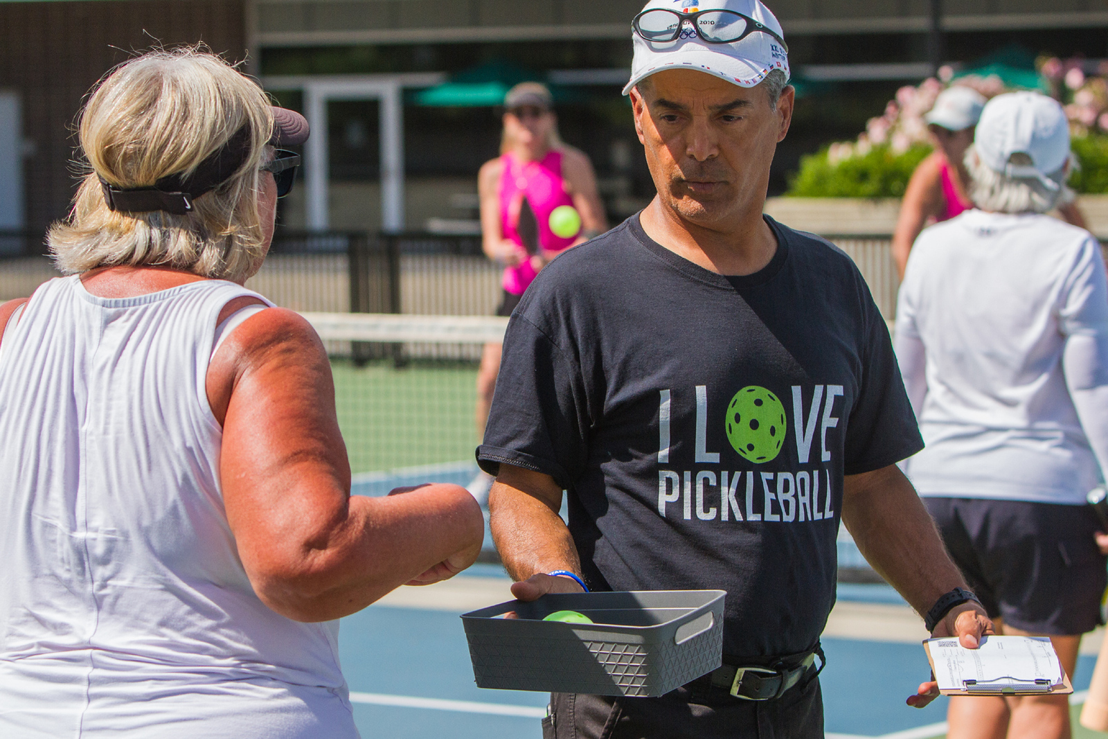 Man in "I love pickleball" shirt holding basket and clipboard talking to woman on outdoor pickleball court