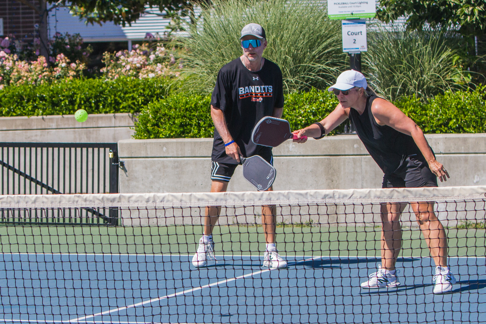 Two adults playing pickleball on an outdoor court, one about to hit a green ball over the net.