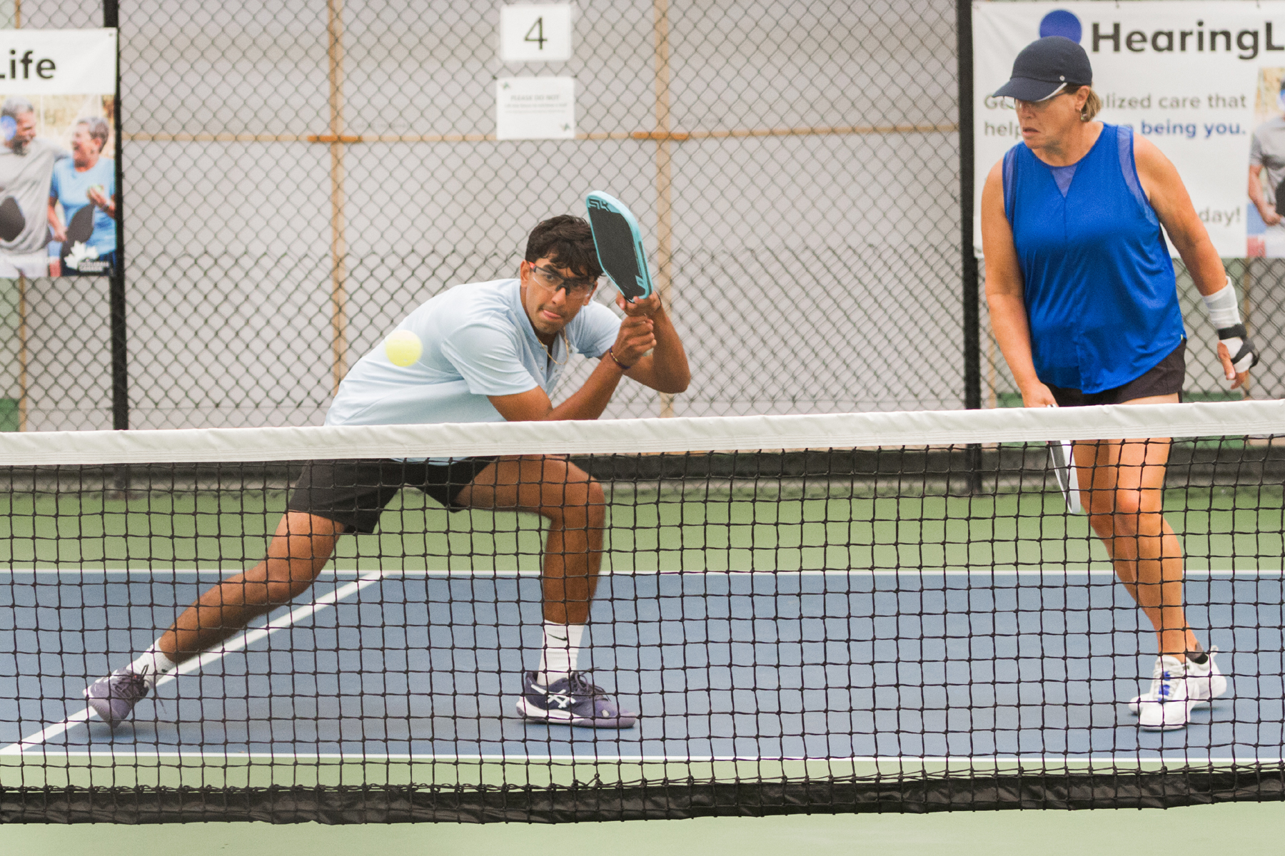 Two pickleball players in action near the net on an indoor court with chain-link fence background