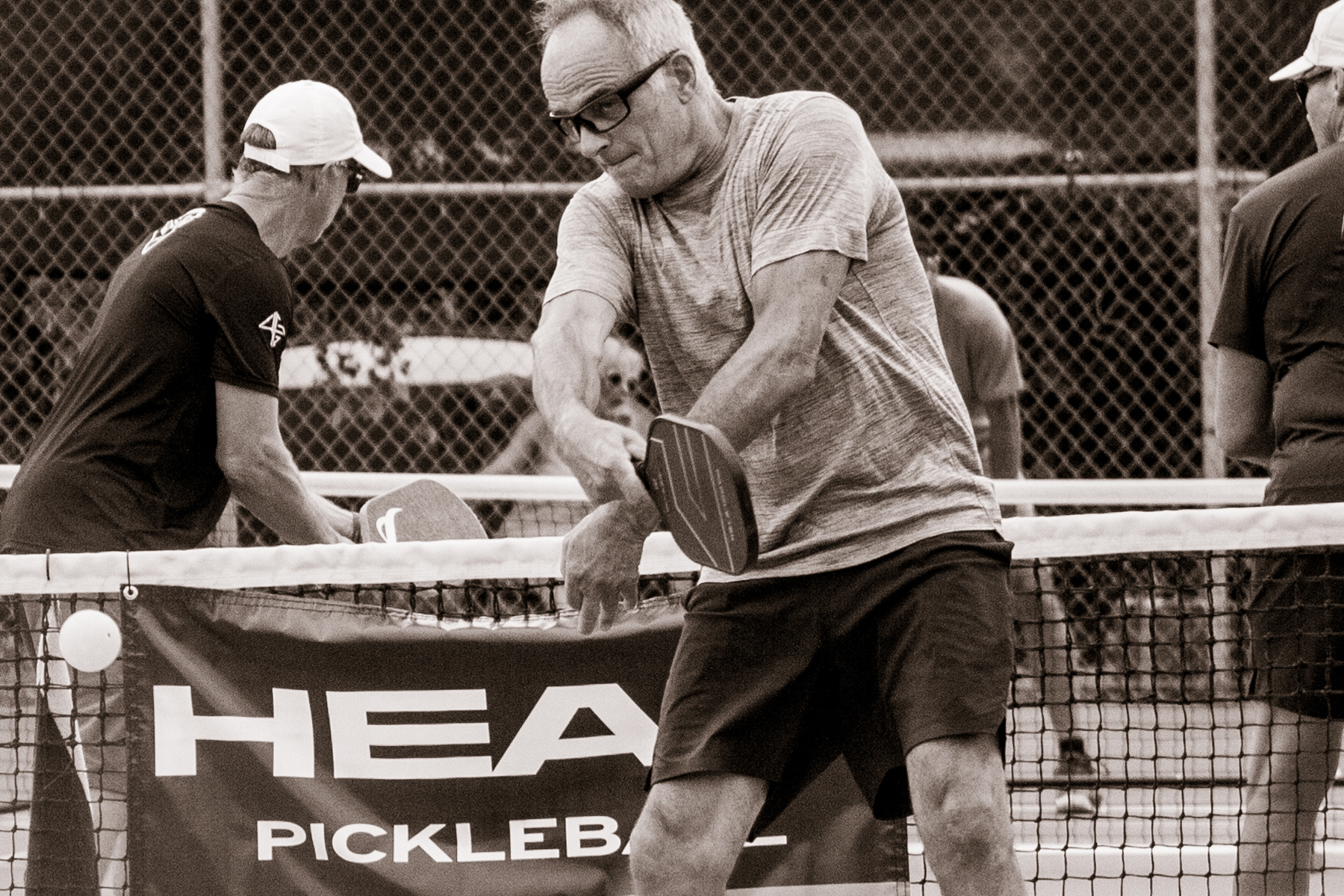 Older man in glasses hitting a pickleball with paddle on outdoor court, two other players visible behind net