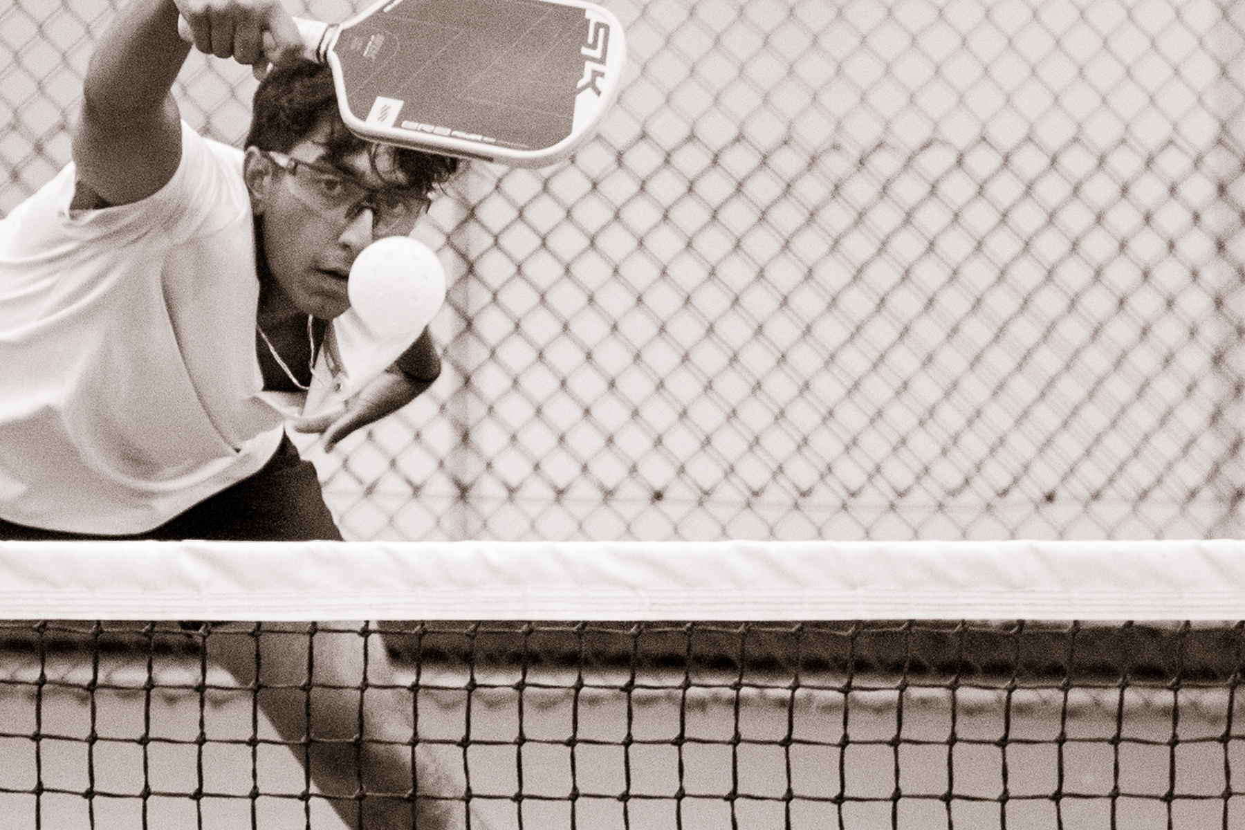 Man wearing glasses hitting a pickleball with a paddle near a net and chain-link fence background