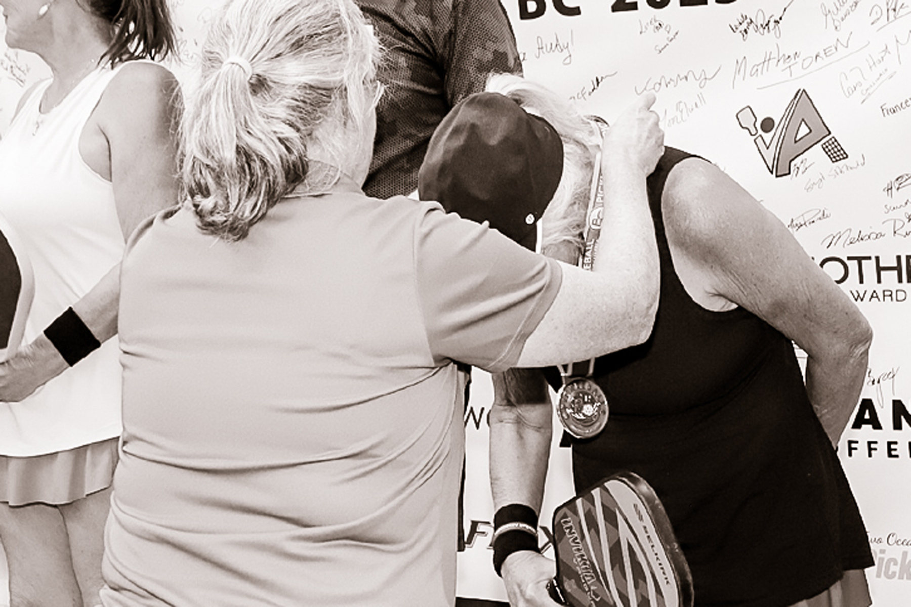 Three women at a pickleball event, one wearing a medal and holding a paddle, signing a large board with signatures.