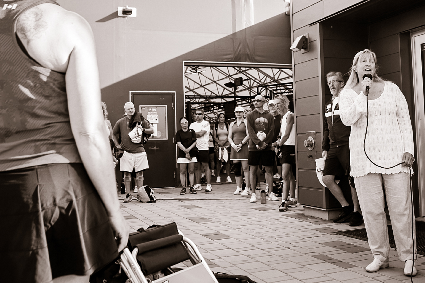 Woman speaking into microphone outside building with group of people standing in line behind her, some in sportswear