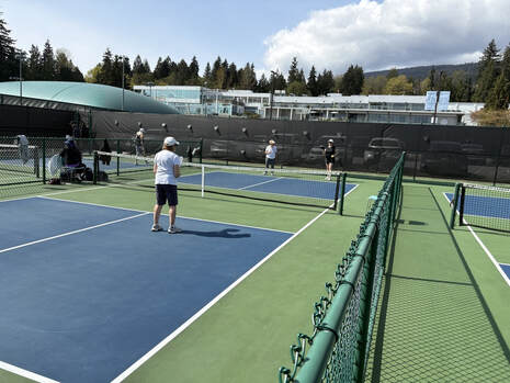 hollyburn-country-club-courts Outdoor pickleball courts with players, surrounded by fencing, trees, and buildings under a partly cloudy sky.