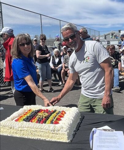 108-mile-scpa-opening Two people cutting a large rectangular cake outdoors with a crowd seated and standing behind them under a blue sky.
