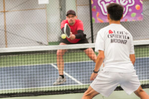 Two men playing pickleball indoors, one in red hitting the ball with a paddle, the other in white ready to return it