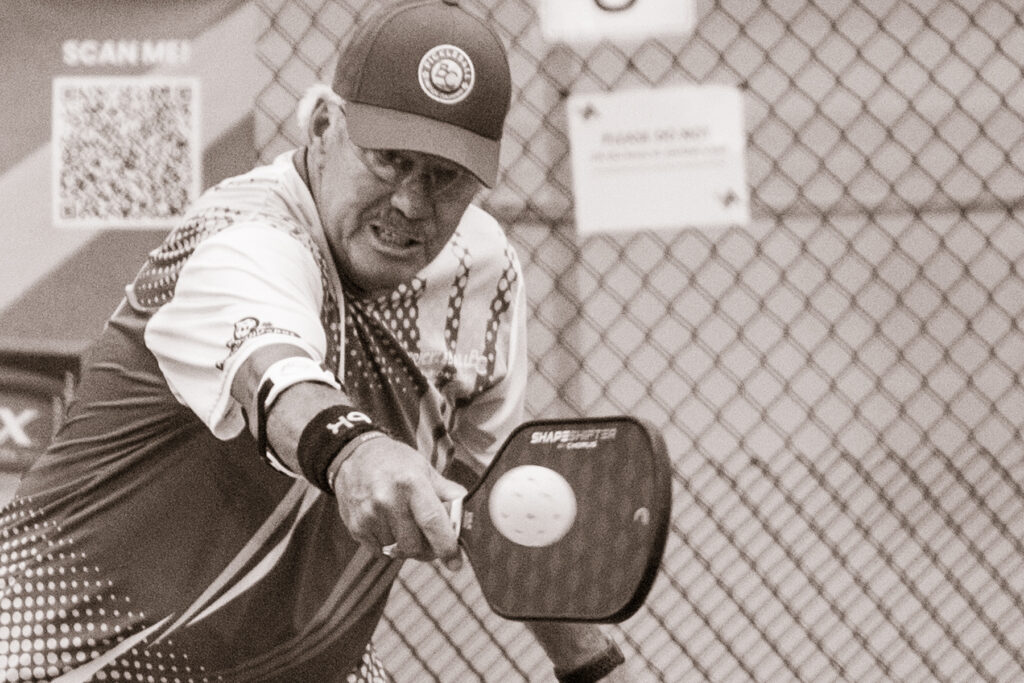 Man wearing cap and sportswear hitting a pickleball with paddle in front of a chain-link fence and QR code sign.