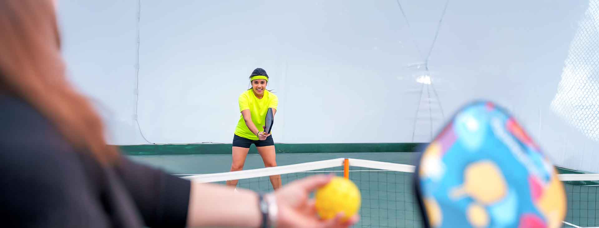 Woman in yellow shirt ready to receive serve in indoor mixed doubles pickleball match with colorful paddle and ball visible