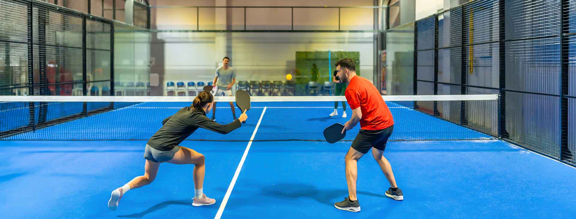 Two people playing pickleball on a blue indoor court with a net and glass walls surrounding the area.