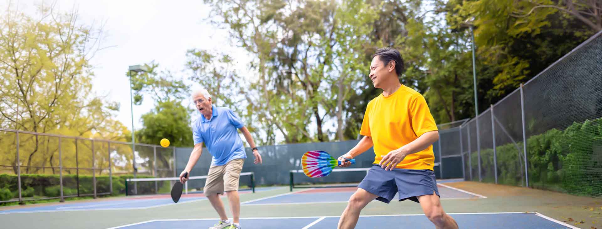 Two men playing outdoor pickleball on a court, one with a rainbow paddle and the other with a black paddle.