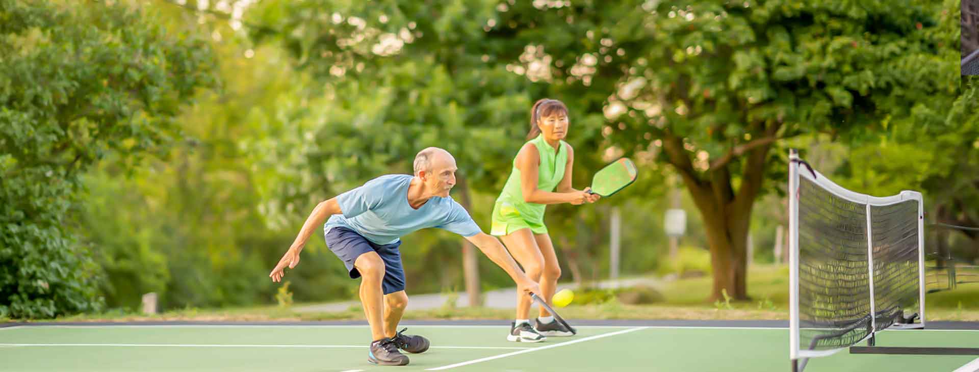 Two seniors playing doubles pickleball outdoors, one reaching low to hit the ball near the net on a green court.