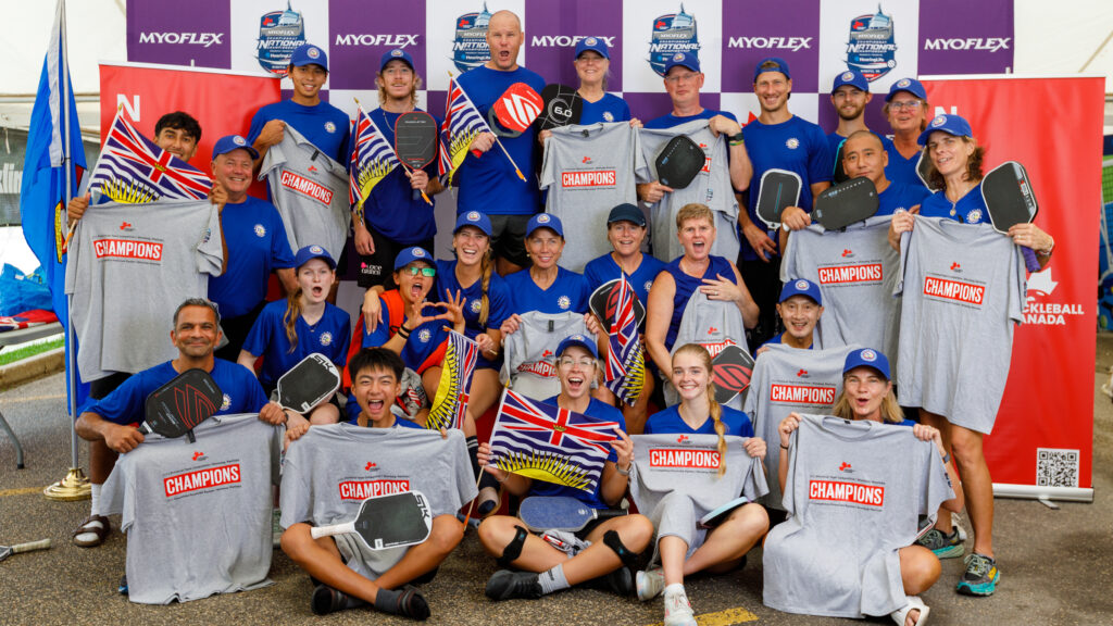 Group of pickleball players in blue shirts holding paddles, BC flags, and gray "Champions" shirts, smiling at camera.